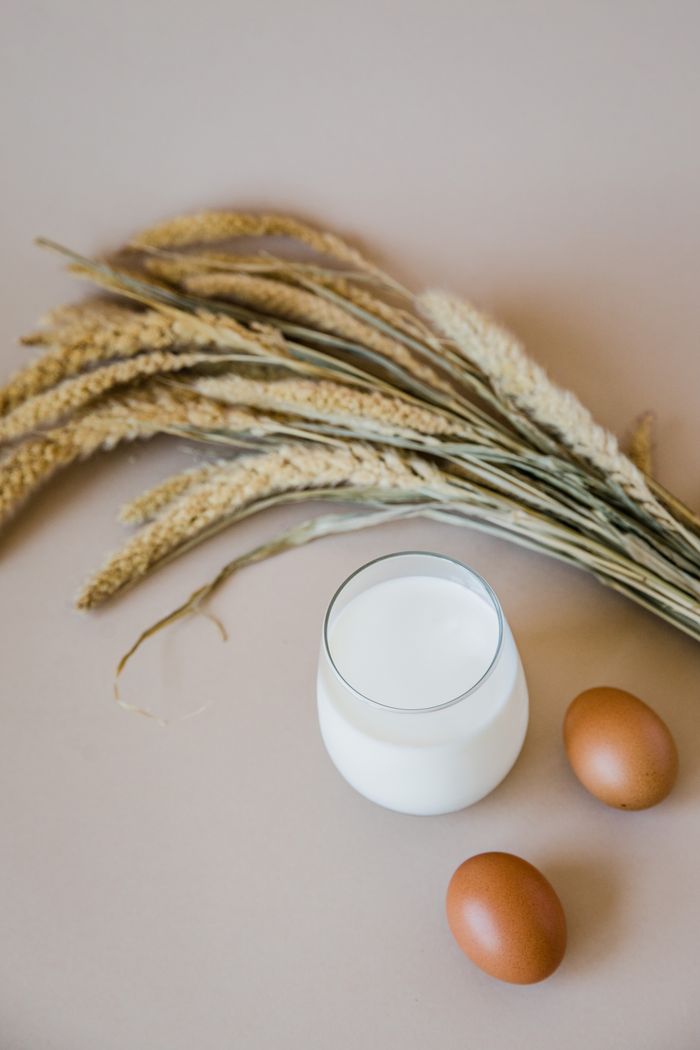 A flat lay of fresh eggs, a glass of milk, and wheat on a neutral background.